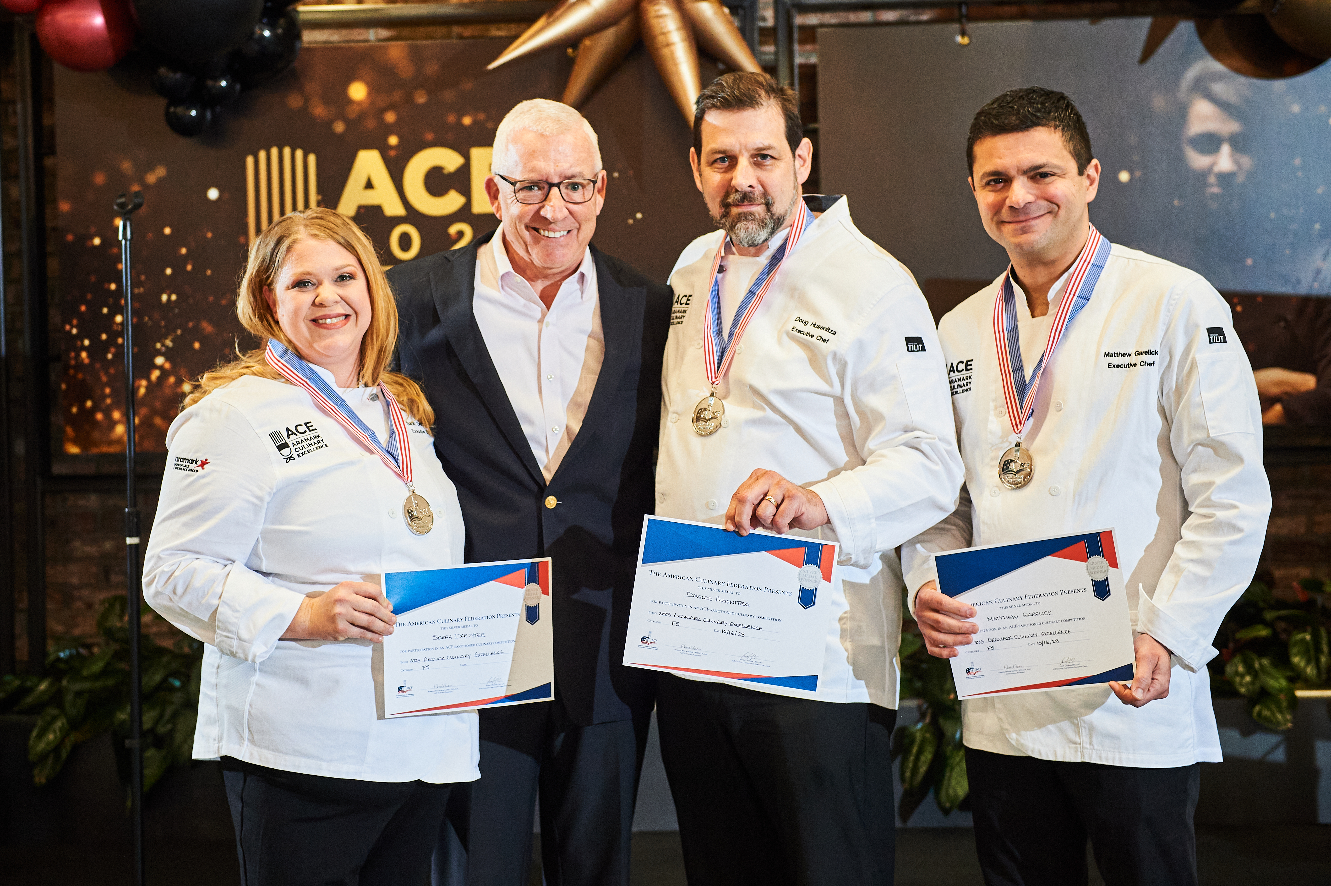 Three chefs and one man in suit smiling with certificates and medals